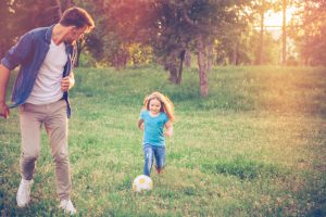 Young father playing soccer ball with daughter at a park. Sun flare, green grass, summer time. Caucasian, blond hair.