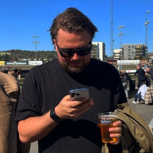 Man i solglasögon och svart t-shirt tittar på sin mobil, med en öl i handen.